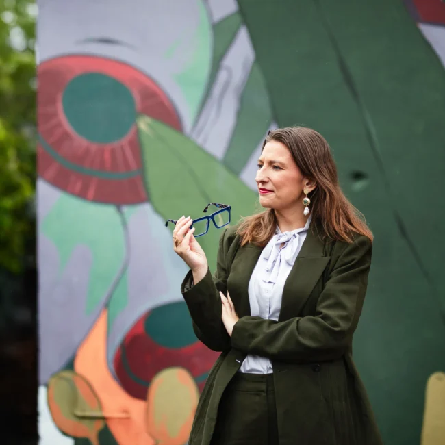 Woman in olive green suit holding glasses in front of a colourful mural.