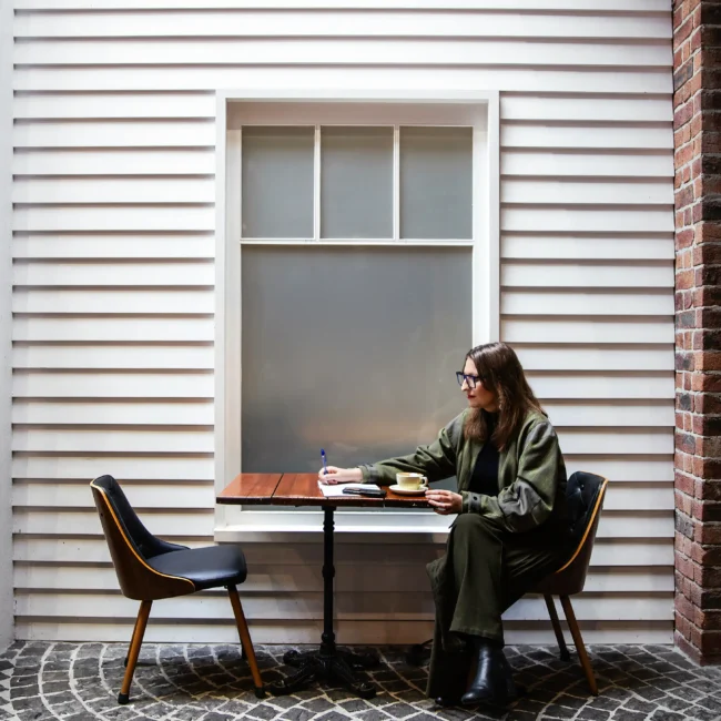 Professional woman sitting at an outdoor cafe table writing on a notepad against a white weatherboard wall.